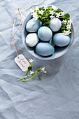 Blue coloured Easter eggs in a ceramic bowl with flower decoration on a blue linen tablecloth