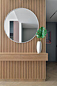 Round mirror above wooden sideboard with monstera leaf in vase