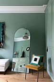 Bedroom with wardrobe and walls in pastel green, full-length mirror and chair with cushions