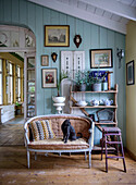 Antique sofa with cat in front of wall with blue wood paneling, shelf with crockery, plants and decorations