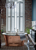 Free-standing copper bathtub in front of window with shutters and brick wall in bathroom