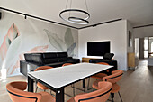 Wohnzimmer mit Wandbild, schwarzer Ledercouch und Marmortisch mit orangefarbenen Stühlen