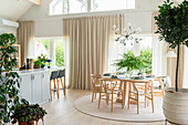 Dining area with round table on round carpet, wooden chairs and indoor plants in bright room