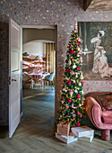 Narrow, decorated Christmas tree next to pink velvet sofa in floral wallpapered living room