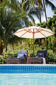 Two sun loungers with umbrella by the pool in front of tropical palm trees