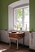 Rustic side table with turned legs and antique chest in front of window with view of greenery