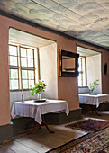 Two tables with white tablecloths and floral arrangements in front of large mullioned windows