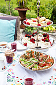 Etagere with sandwiches, mini pavlovas and scones in the garden, tablecloth with floral pattern