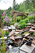 Garten mit Wasserspiel, Holzpergola und üppiger Bepflanzung im Sommer