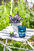 Blue glass with crocheted DIY decoration, horned violets and forget-me-nots in a flower pot on a white wooden chair in the garden