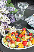 Obstplatte mit Erdbeeren, Melonen und Trauben neben Fliederstrauß auf Tisch