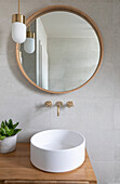 Round mirror with wooden frame above modern washbasin with golden fittings