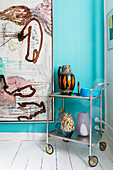 Serving trolley with vintage jugs and vases in front of an abstract painting in a turquoise-coloured room