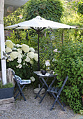 Small garden alcove with bamboo umbrella and hydrangeas, anthracite-coloured folding furniture on gravel path