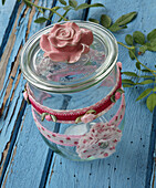 Self-sewn ribbons made of rose fabric, mini roses and fabric rosettes as decoration for storage jar and glass lid with rose on blue wooden table
