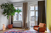 Living room with large rubber tree, retro armchair, heavy curtains and balcony access