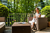 Frau mit Weinglas sitzt auf einem Balkon mit Loungemöbeln, Hamburg, Deutschland