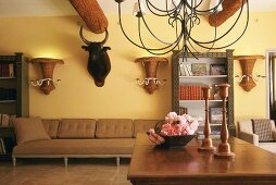 Wohnzimmer mit imposanten Balken an der Decke, Stierkopf, vasenförmigen Korblampen an der Wand und filigraner, eiserner Hängelampe über Holztisch mit Blumendekoration
