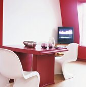 Ein roter Esstisch mit weissen Panton Stühlen in einer Zimmerecke mit leicht schräger Wand