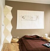 Ein einfaches Bett mit Tagesdecke zwischen zwei Bodentischen aus Rattan, an der braunen Wand eine abstrakte Zeichnung