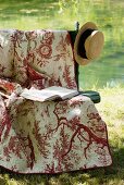 Romantische Gartenbank mit Decke, Buch und Strohhut am Teich