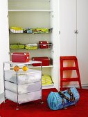 A rolling cabinet, a bag and a red step stool in front of a wardrobe with an open door