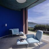 A living room with upholstered loungers in front of a curved, blue-painted wall and a view of the sea