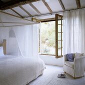 A white bedroom in a country house - a four poster bed covered with light fabric in front of an open window
