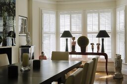 A dining table in front of an elegant occasional table set with table lamps in a bay window with closed shutters