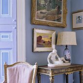 A sculpture and a table lamp with a white shade on an antique wall table and pictures hanging on a lilac-painted wall