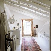 Mediterraner Bad unter dem Dach mit weiss gestrichenen Holzbalken und Teppichläufer vor Fenster mit drapiertem Vorhangschal