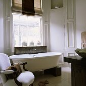 Free standing bathtub on wooden blocks and an antique white chair in front of window in a light gray bathroom