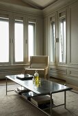 Couchtisch mit Metallgestelll und Sessel in Wohnraumecke mit hellgrauer Vertäfelung und Fenstern