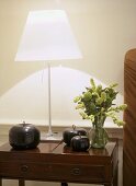 A detail of a modern sitting room showing a polished wood side table, lit steel lamp with white shade, ornaments, flower arrangement