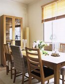 A modern dining room in neutral colours, wooden dining table, chairs, window, venetian blinds, display cabinet, vase, flowers,
