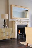 Lamp on chest of drawers next to traditional style fireplace with lit fire in contemporary sitting room