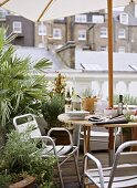 A detail of a roof garden patio terrace, a wood table with parasol, metal chairs, plants in containers