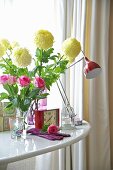 A bunch of flowers in a glass vase and an alarm clock in front of a desk lamp in a white table