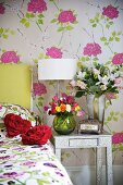 Various bunches of flowers on a bedside table against a wall with floral patterned wallpaper