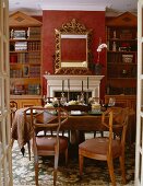 Blick in ein rotes Esszimmer mit hohen Bücherregalen auf beiden Seiten des Kamins, darüberhängendem Spiegel mit Goldrahmen und Biedermeierstühlen