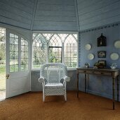 Weisser Rattansessel auf Sisalteppich vor gotischen Fenstern in pastellblauem Holzpavillon