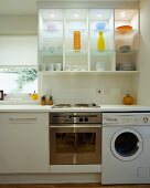 Ein Chrombackofen und eine weiße Waschmaschine stehen unter einer beleuchteten Vitrine aus Holz und Glas, die mit farbigen Vasen und Geschirr eingedeckt wurde