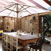 In einem Wintergarten, der gleichzeitig als Speisesaal dient, bietet ein weißer Sonnenschirm einem alten Kiefer-Tisch mit Stühlen Schatten