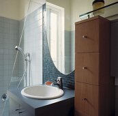 Ein rundes Waschbecken ist in einem grauen Unterschrank eingebracht worden, der zwischen eines hohen Schubladenschrank und einer Dusche steht