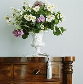 Nahaufnahme eines weiss-violetten Blumenstrausses in weisser Amphorenvase auf antiker Holzkommode