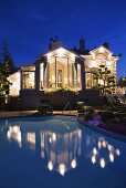 Hell beleuchtetes Haus mit großflächigem Pool und sich spiegelnden Lichtern auf der Wasseroberfläche