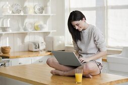 A young woman with laptop sitting on a kitchen counter
