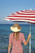 Frau mit Hut und rot-weißem Sonnenschirm am Meer