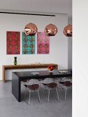 Colourful, graphic artworks behind retro-style dining area with wire chairs, console table and spherical, metallic lamps