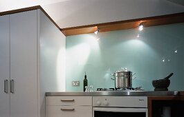 A white, modern kitchen with a glass splashback and spotlights built into the underside of a wooden shelf